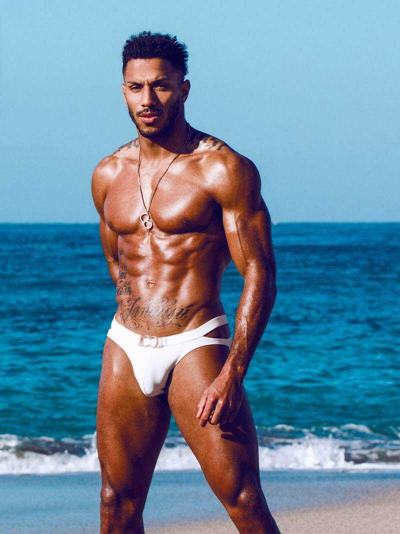 Muscular man in white swim trunks standing on a beach with blue ocean and sky.