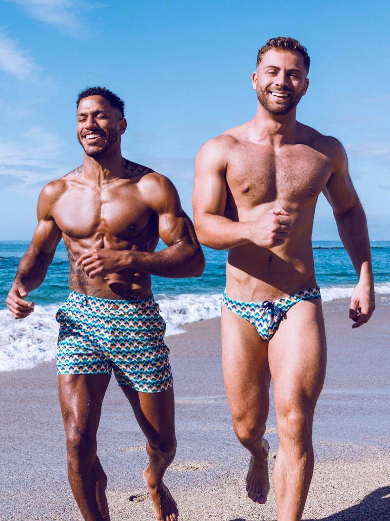 Two men running on a beach wearing patterned swim trunks.