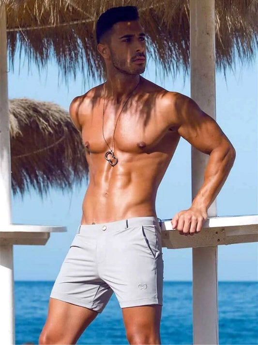 Muscular man in light gray shorts standing by a beach with clear blue sky and ocean.