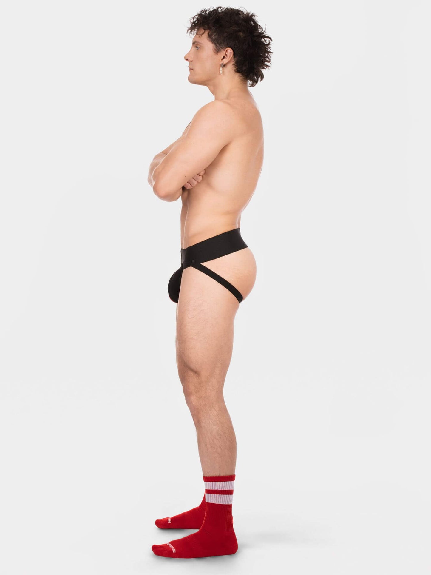 A side-profile studio photo of a person standing against a light gray background, wearing a minimal black strappy underwear-style garment and bright red socks with white stripes.