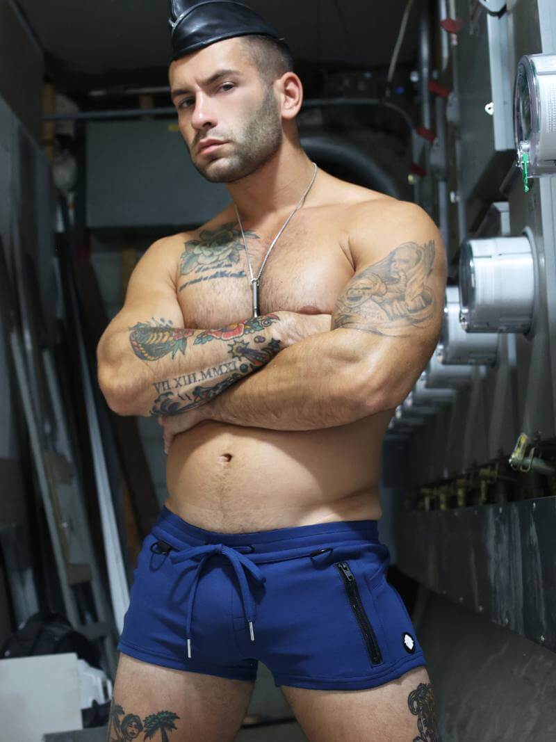 A person with tattoos wearing navy blue mini shorts with a drawstring, featuring a low-rise waist and super short legs, in a mechanical setting.