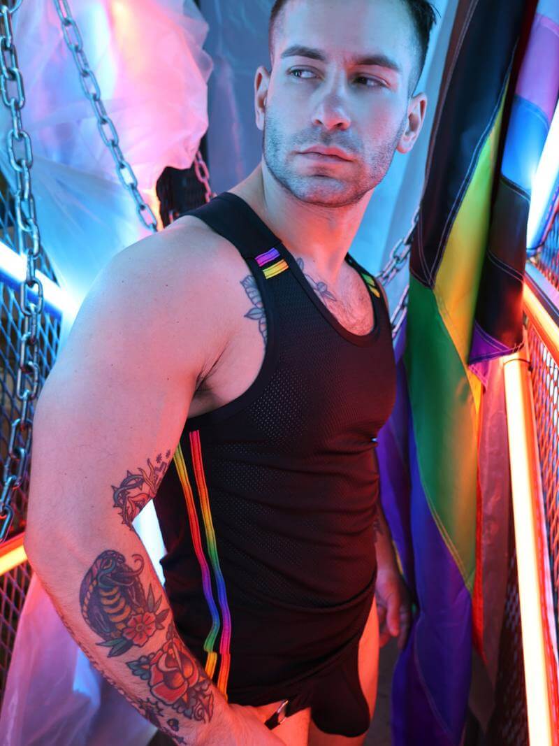 Man in a black tank top with tattoos standing in front of a colorful flag.