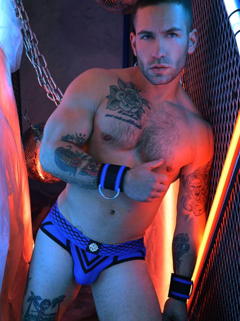 Man wearing blue and black underwear with tattoos on his arms and chest, standing in a dimly lit room.