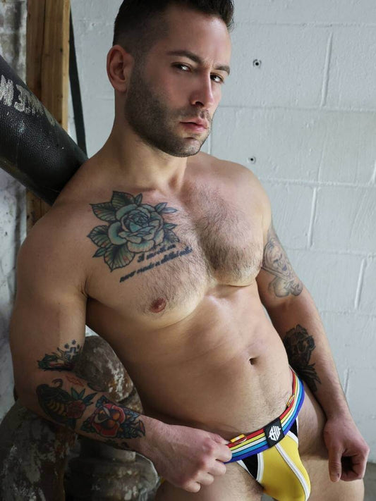 Man with tattoos wearing colorful underwear against a white brick wall.