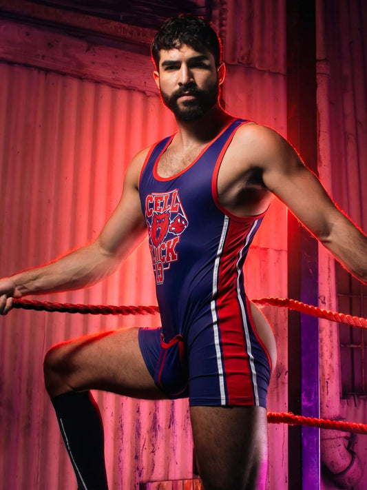 A bearded man wearing a blue, red, and white wrestling singlet stands inside a wrestling ring, posed with one leg raised. The background features a rustic, red-lit corrugated metal wall.