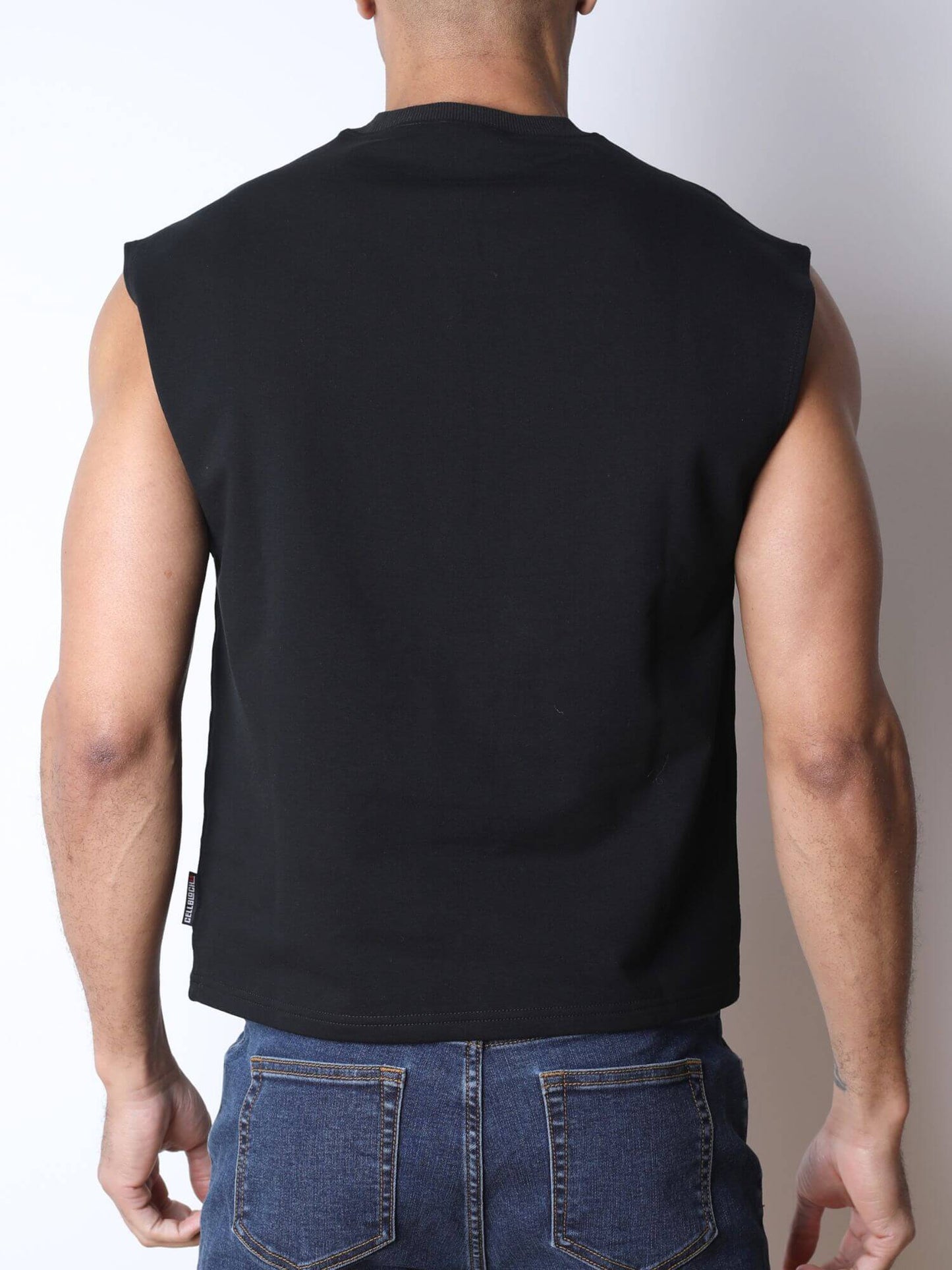 Rear view of a man wearing a black sleeveless muscle shirt and blue jeans against a plain light gray background.