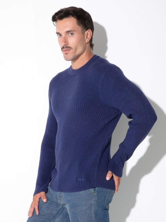 A man wearing a navy blue ribbed knit sweater and blue jeans, standing against a white wall with a soft shadow.