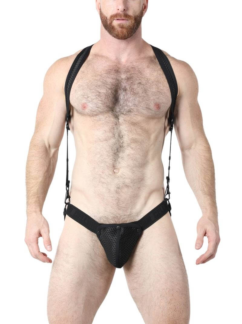 A model wearing a black mesh body harness and jockstrap set with green trim and black metallic hardware. The harness is adjustable with buckles and snaps.