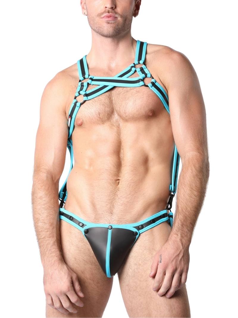 A model wearing a black neoprene body harness with double cross straps across the chest and a jockstrap. The harness has metallic rings and buckles, and the pouch is detachable.