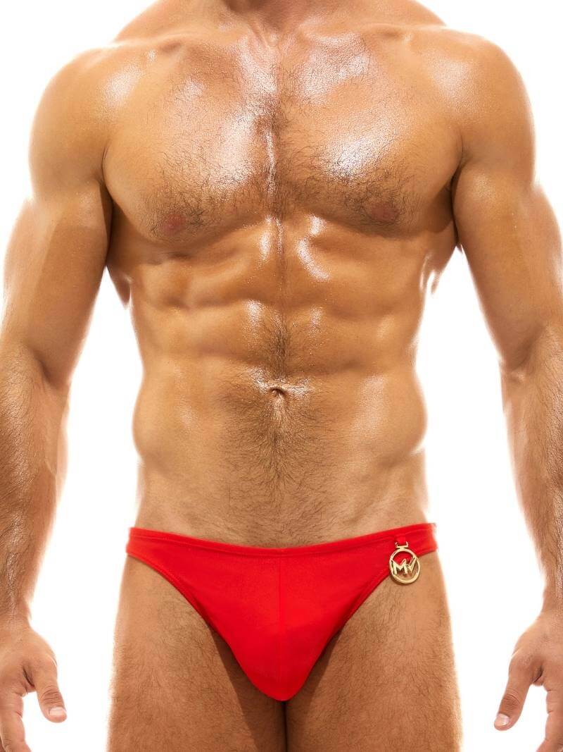 This image features a male model sporting the MODUS VIVENDI Glam Meander Luxury Swim Bikini in gold/silver foil print. The swimwear is designed with vibrant and bold colors, adhering to a luxurious aesthetic with the addition of metallic gold or silver charms and the Meander Greek Key pattern on the rear.