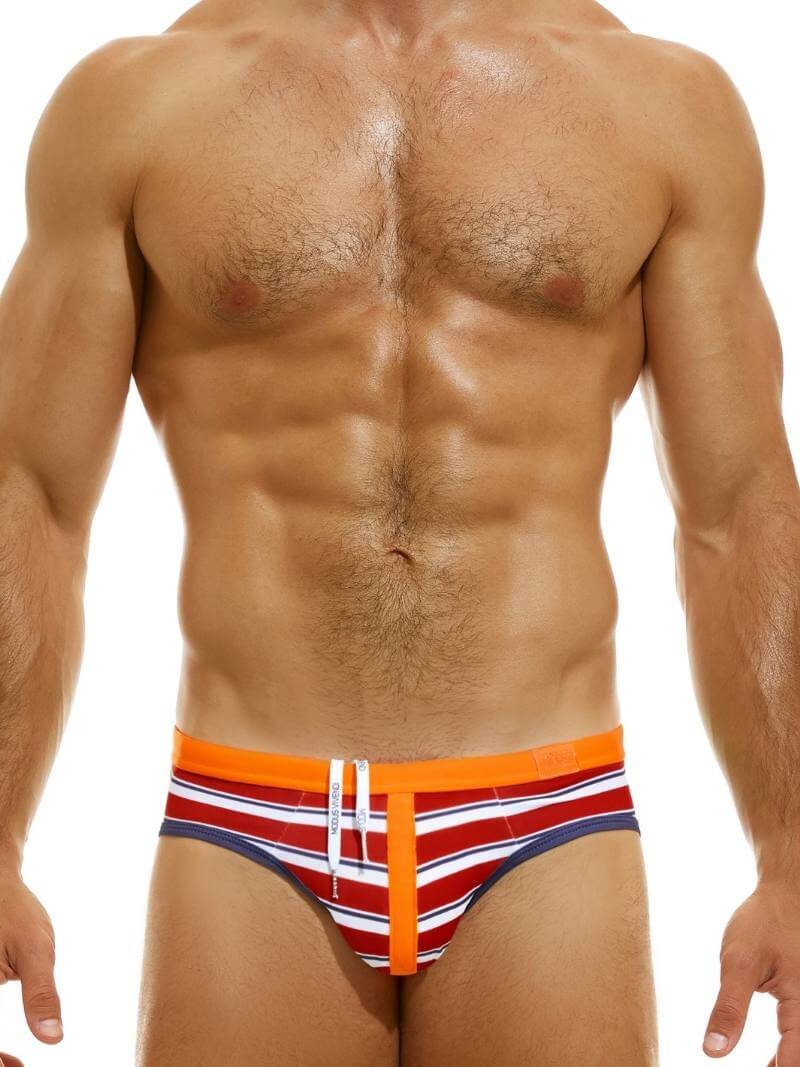 This image features the MODUS VIVENDI Marine Striped Swim Brief, modeled to showcase its fit and style. The swim brief has a striking design with bold, nautical-inspired stripes in red and blue, accented by fluorescent green details.
