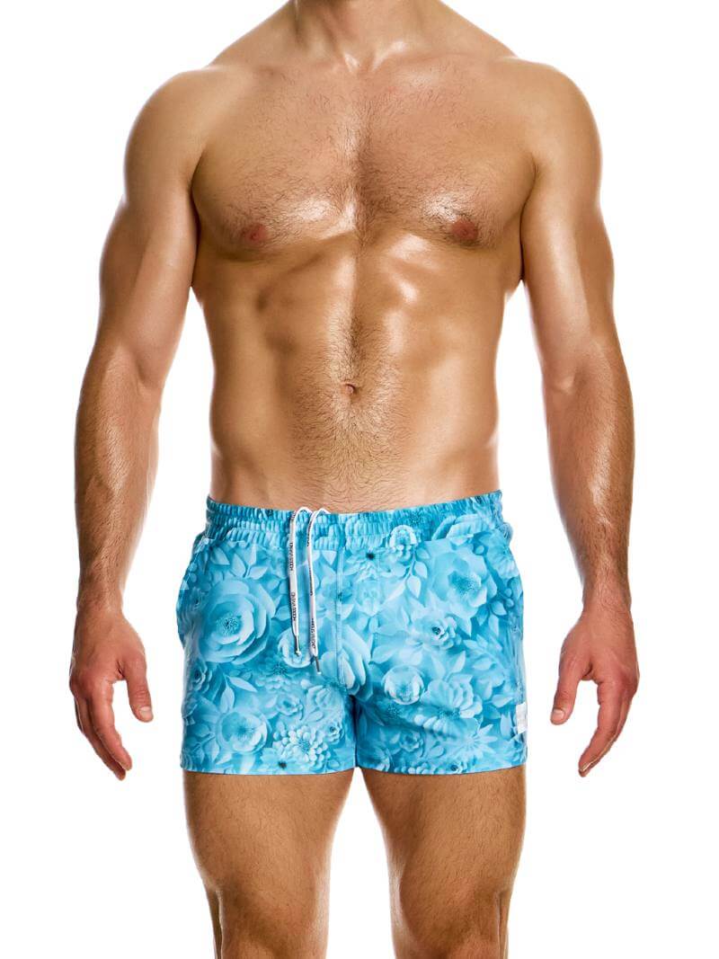 A pair of floral origami swim shorts by Modus Vivendi, featuring a short leg design with pockets, in a pink and blue colorway, modeled by a male swimwear model.