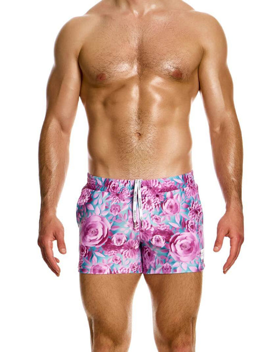 A pair of floral origami swim shorts by Modus Vivendi, featuring a short leg design with pockets, in a pink and blue colorway, modeled by a male swimwear model.