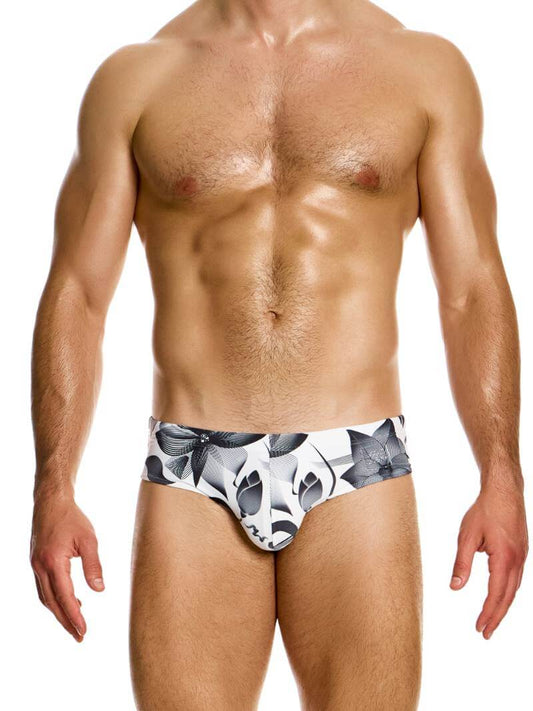 A male model wearing white swim briefs with a black floral print design, featuring an elastic waistband with a Modus Vivendi transparent brand label, and internal drawstrings.