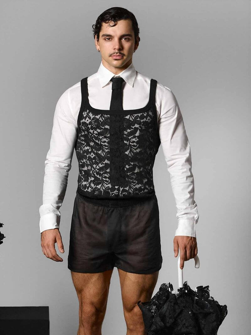 Man wearing a black lace vest over a white shirt with dark shorts, holding a black umbrella on a gray background.