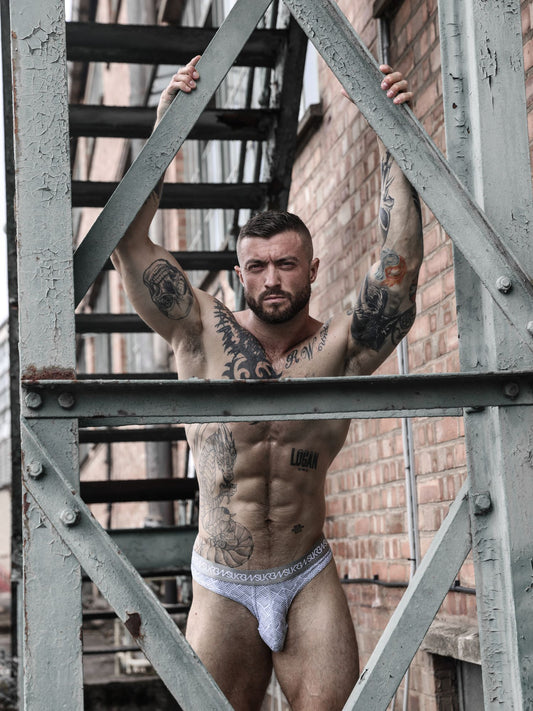 A muscular tattooed man wearing grey briefs stands behind a weathered metal fire escape in front of a brick building.