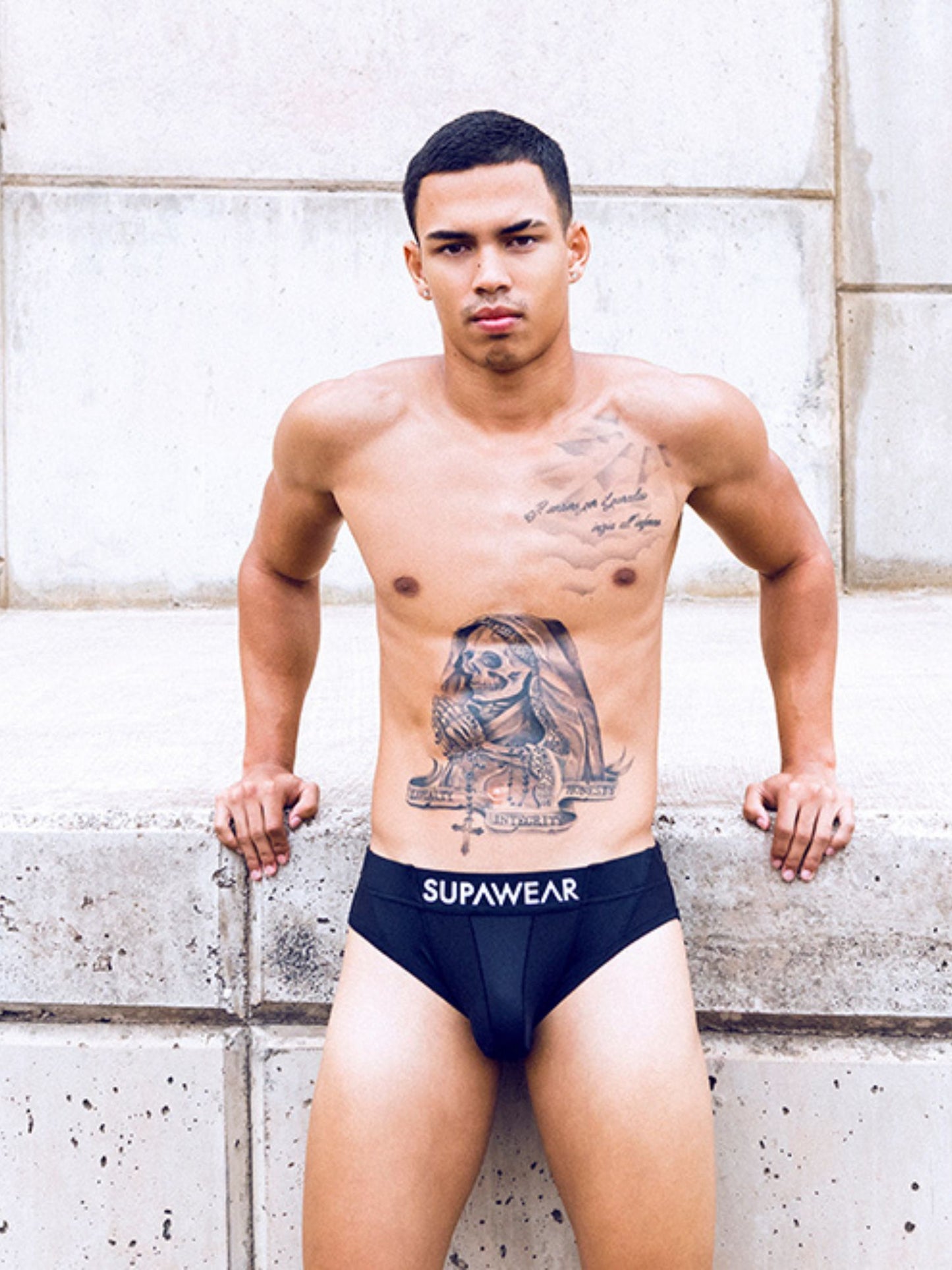 Man wearing black underwear with 'Supawear' branding against a stone wall.