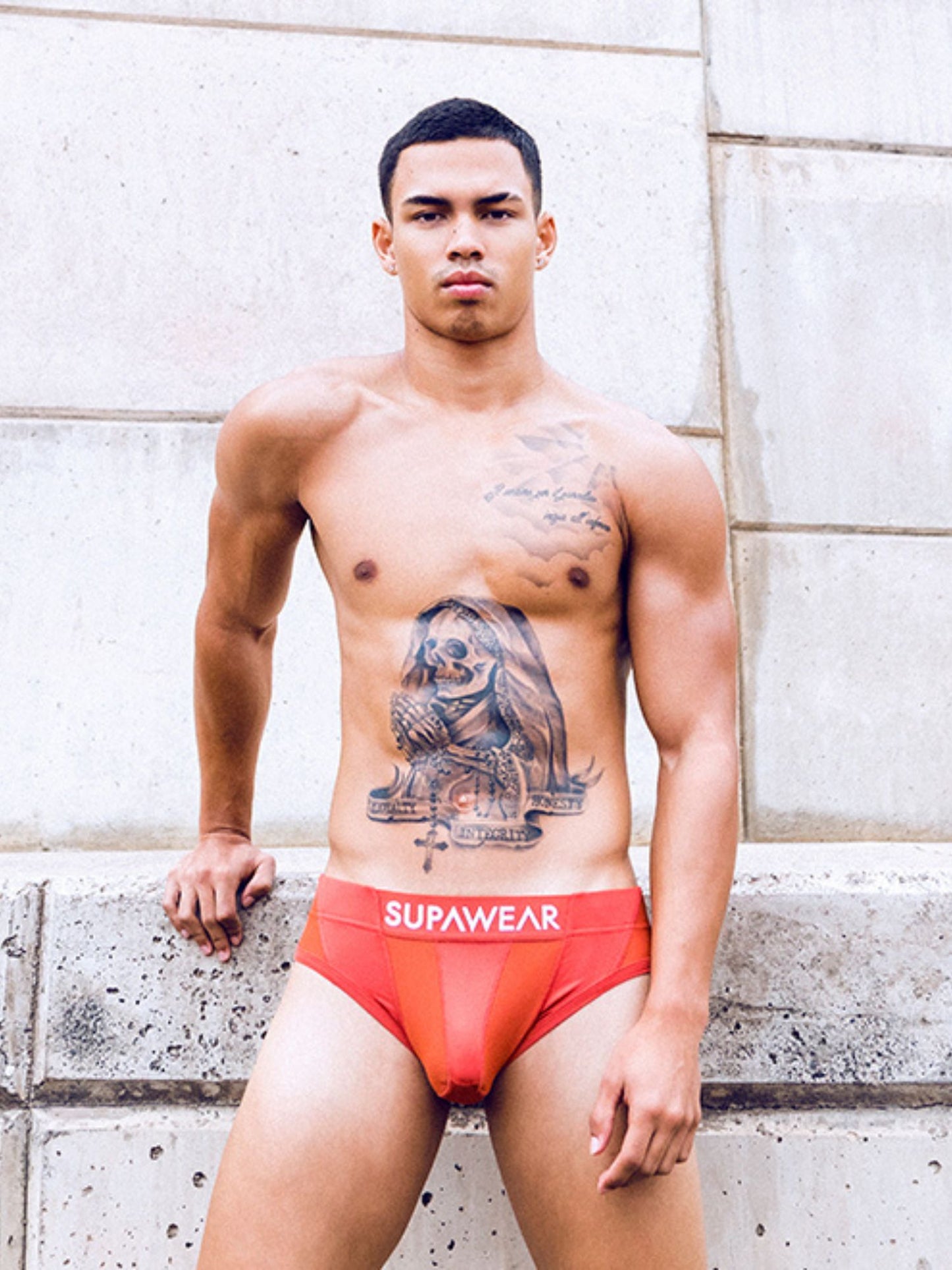 Man wearing red underwear with 'Supawear' logo against a stone wall.