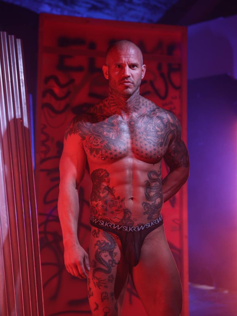 Man with tattoos standing in a room with red lighting and graffiti on the wall.