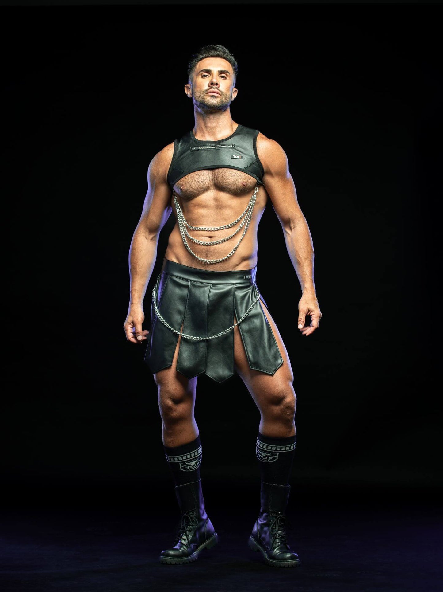 A muscular man modeling a black leather-style costume set consisting of a sleeveless top, matching skirt, and layered chain details, standing against a solid black background.