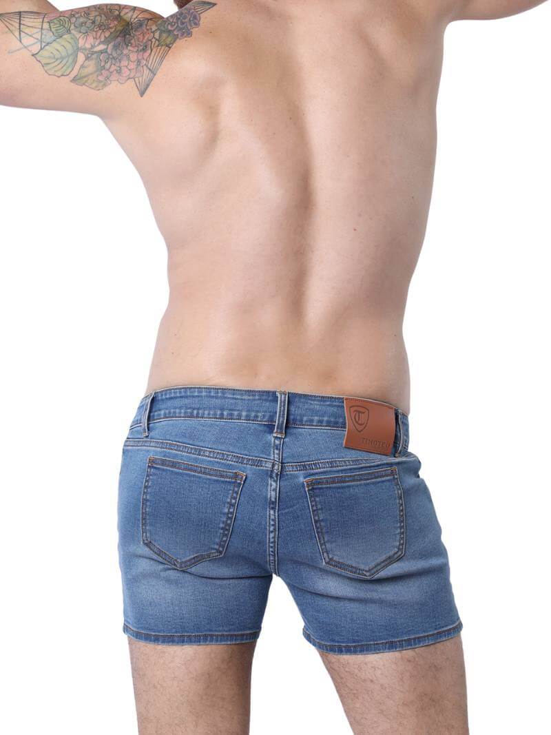 A pair of denim booty shorts with a low-rise waist and a 4-inch inseam, featuring a zipper fly, button fastening, belt loops, and four pockets. The shorts are blue with contrast stitching, and there is a leather branded patch on the back.
