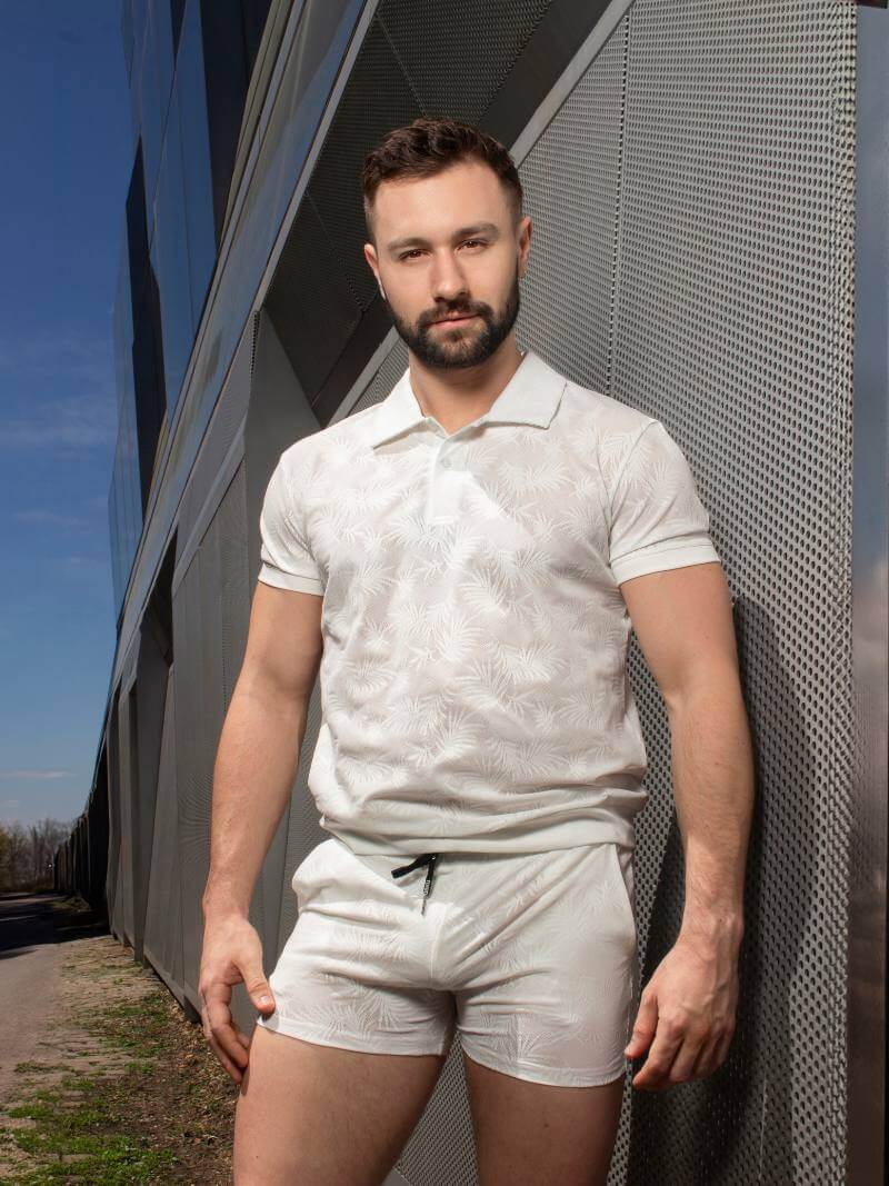 Man wearing a white patterned polo shirt and shorts standing against a modern building.