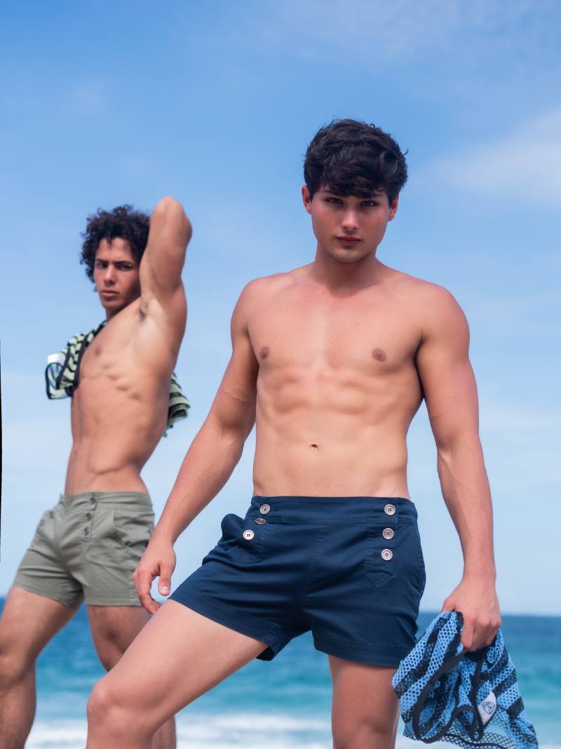 Two men standing on a beach wearing swim trunks with a clear blue sky in the background.