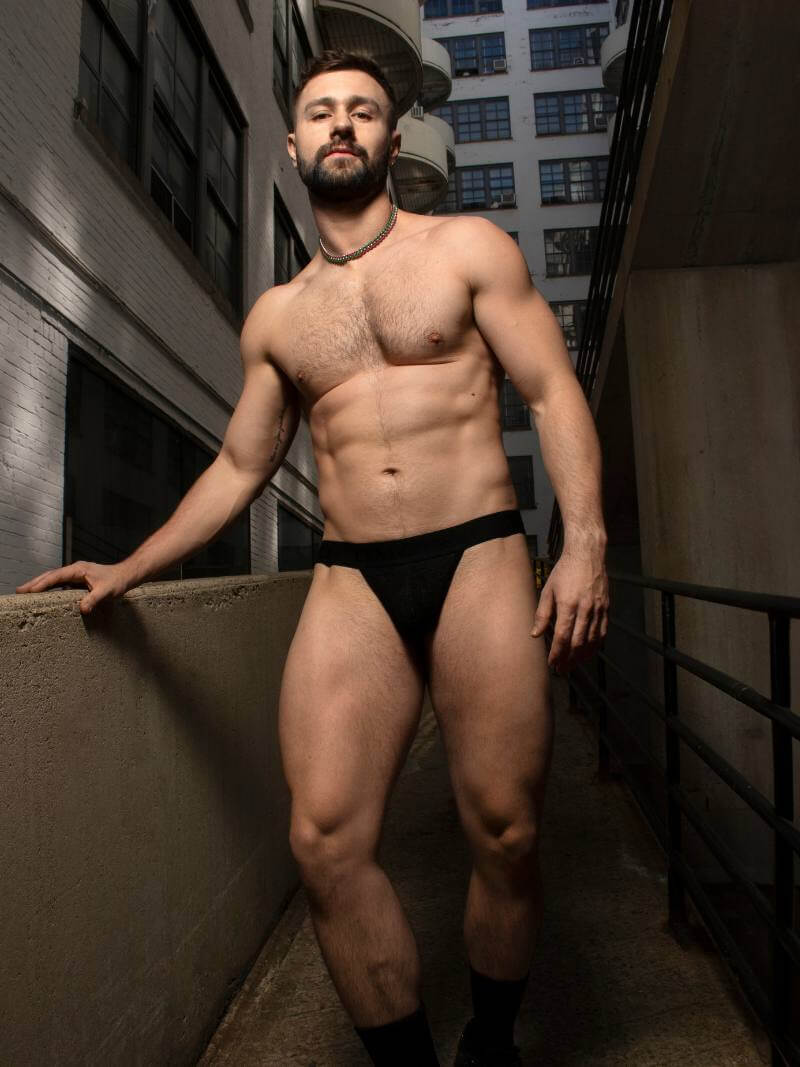 Man wearing a black thong standing in an urban alleyway.