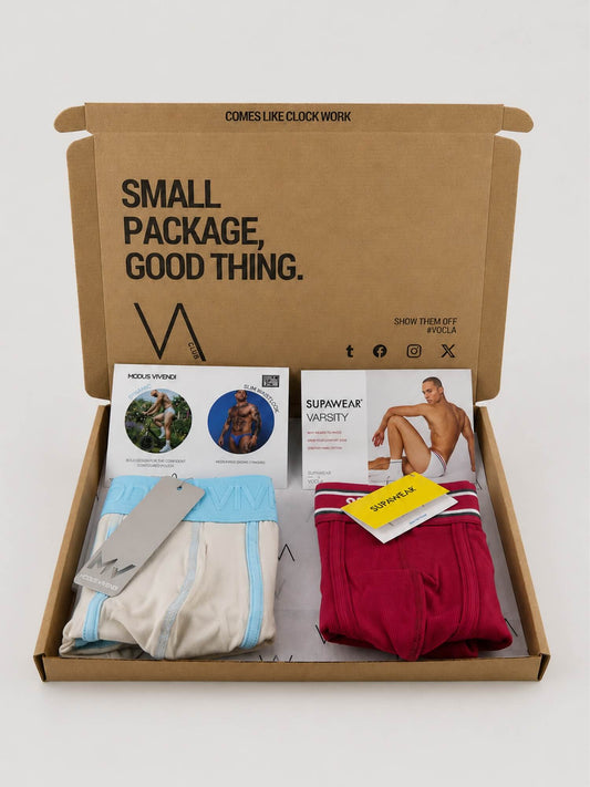 An open brown cardboard gift box on a light gray background, containing folded underwear and printed inserts. Visible items include light blue and off-white underwear, a folded pair of dark red underwear with white stripes, a gray tag, and printed cards reading 'SMALL PACKAGE, GOOD THING.