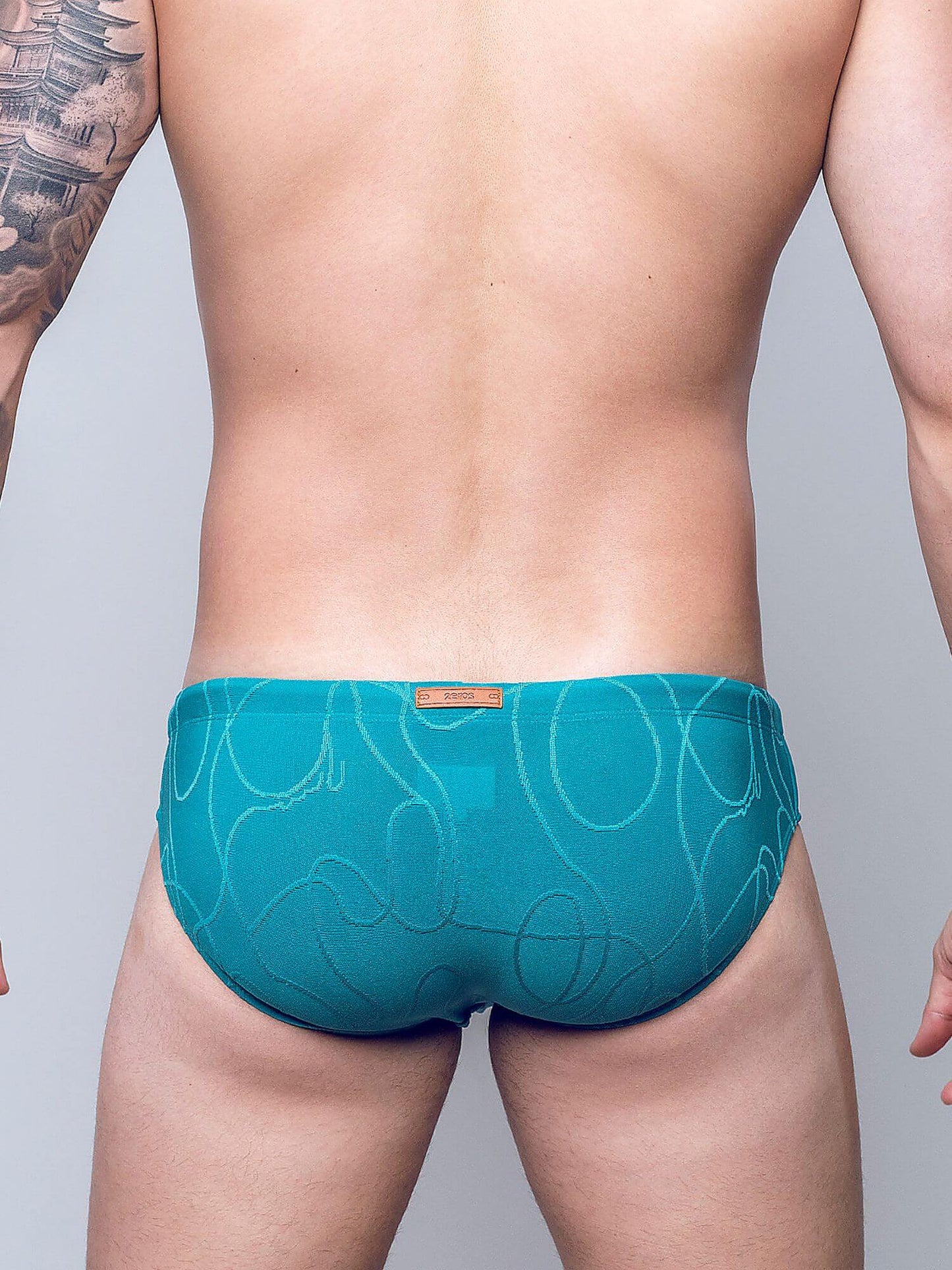 Rear view of a man wearing teal blue patterned briefs with a small tan brand tab at the center back waistband, against a light gray studio background.