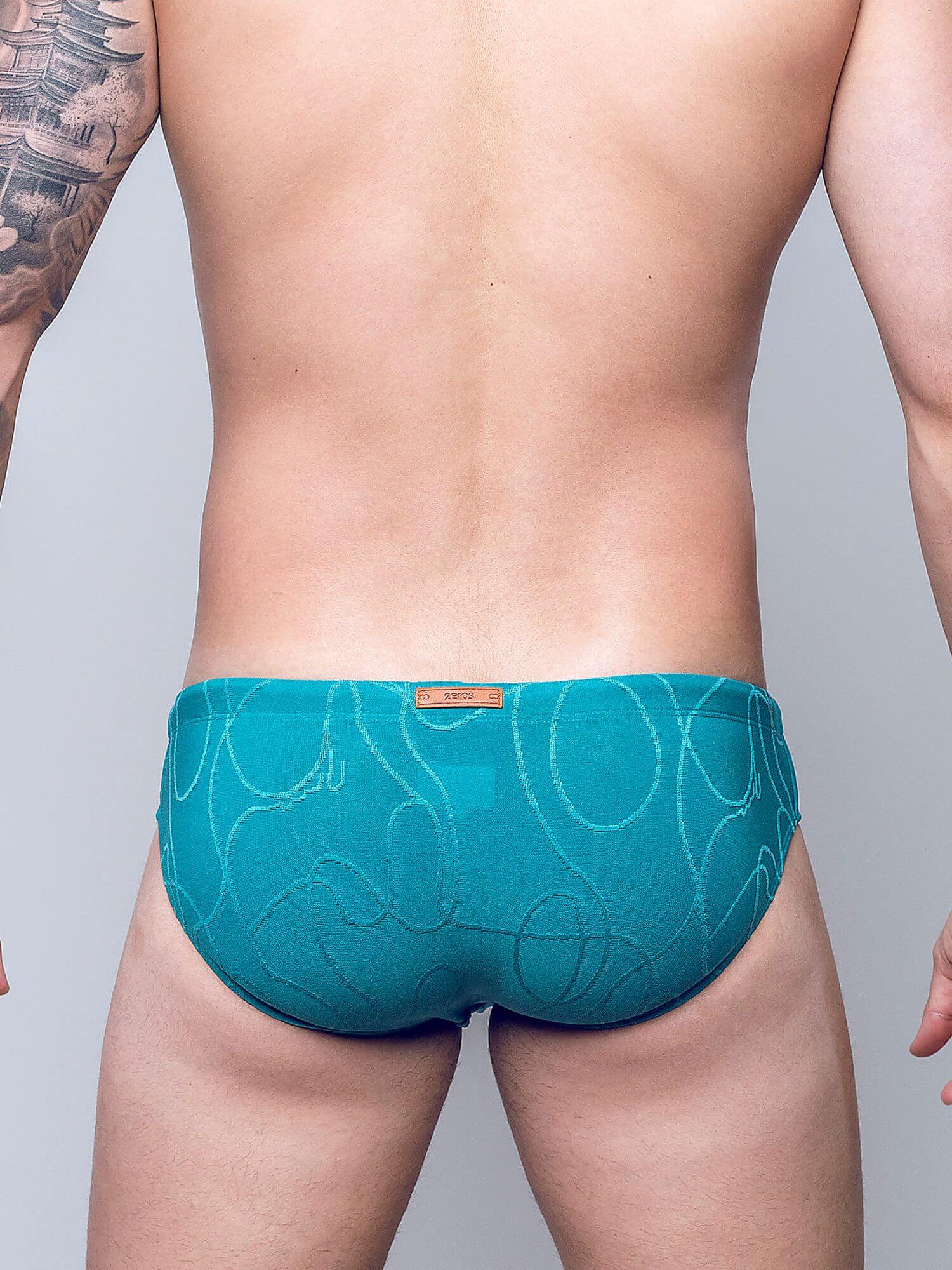 Rear view of a man wearing teal blue patterned briefs with a small tan brand tab at the center back waistband, against a light gray studio background.