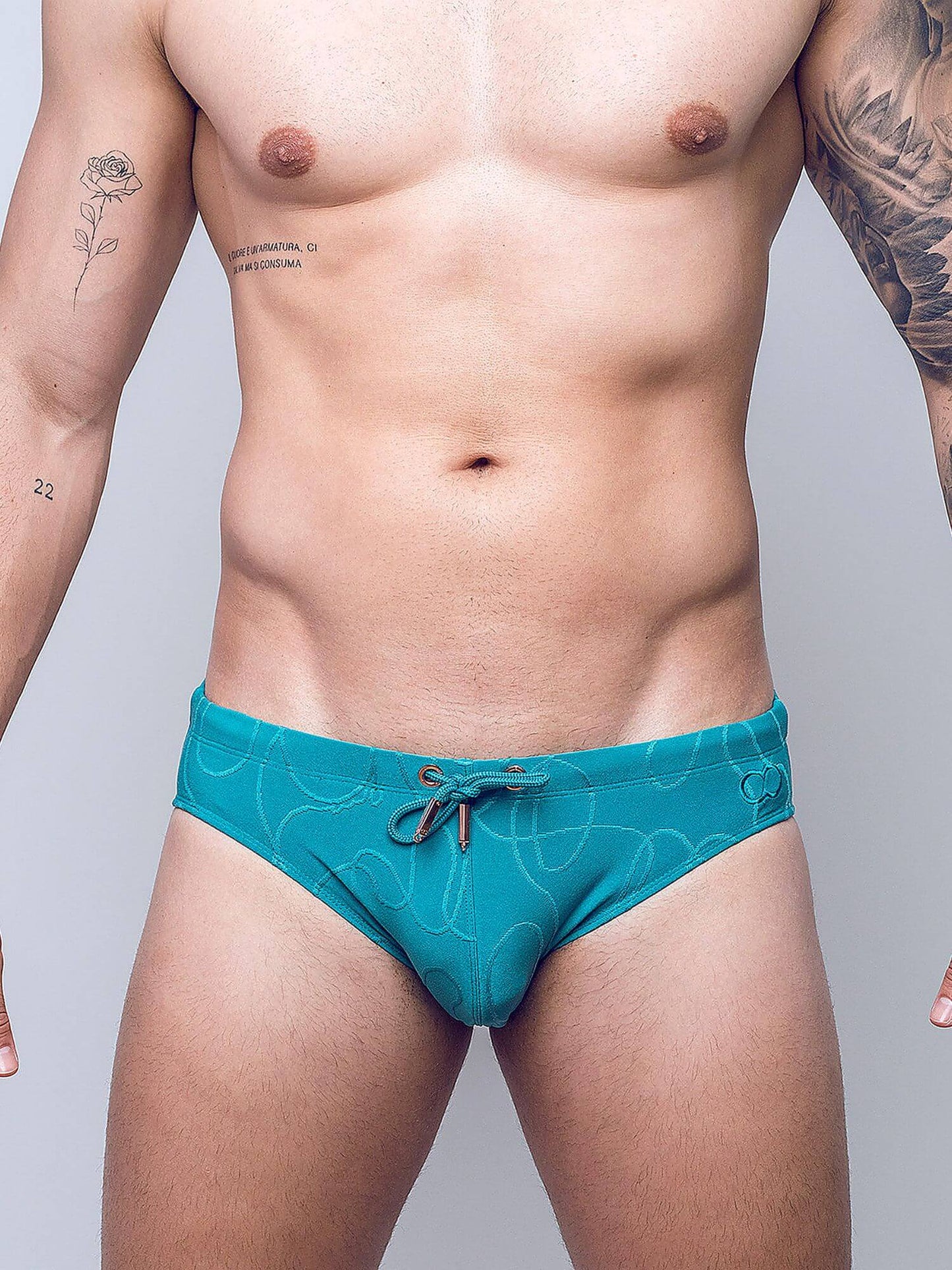 A close-up of a man's torso wearing teal men's swim briefs with a subtle white line pattern and a small brown drawstring, photographed against a light gray studio background.