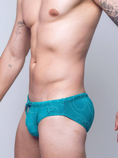 A close-up of a man's torso wearing teal men's swim briefs with a white wavy line pattern, set against a plain light gray studio background.