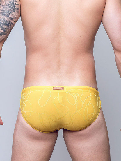 Rear view of a man wearing bright yellow men's briefs with a subtle white abstract line pattern, set against a plain light gray studio background.