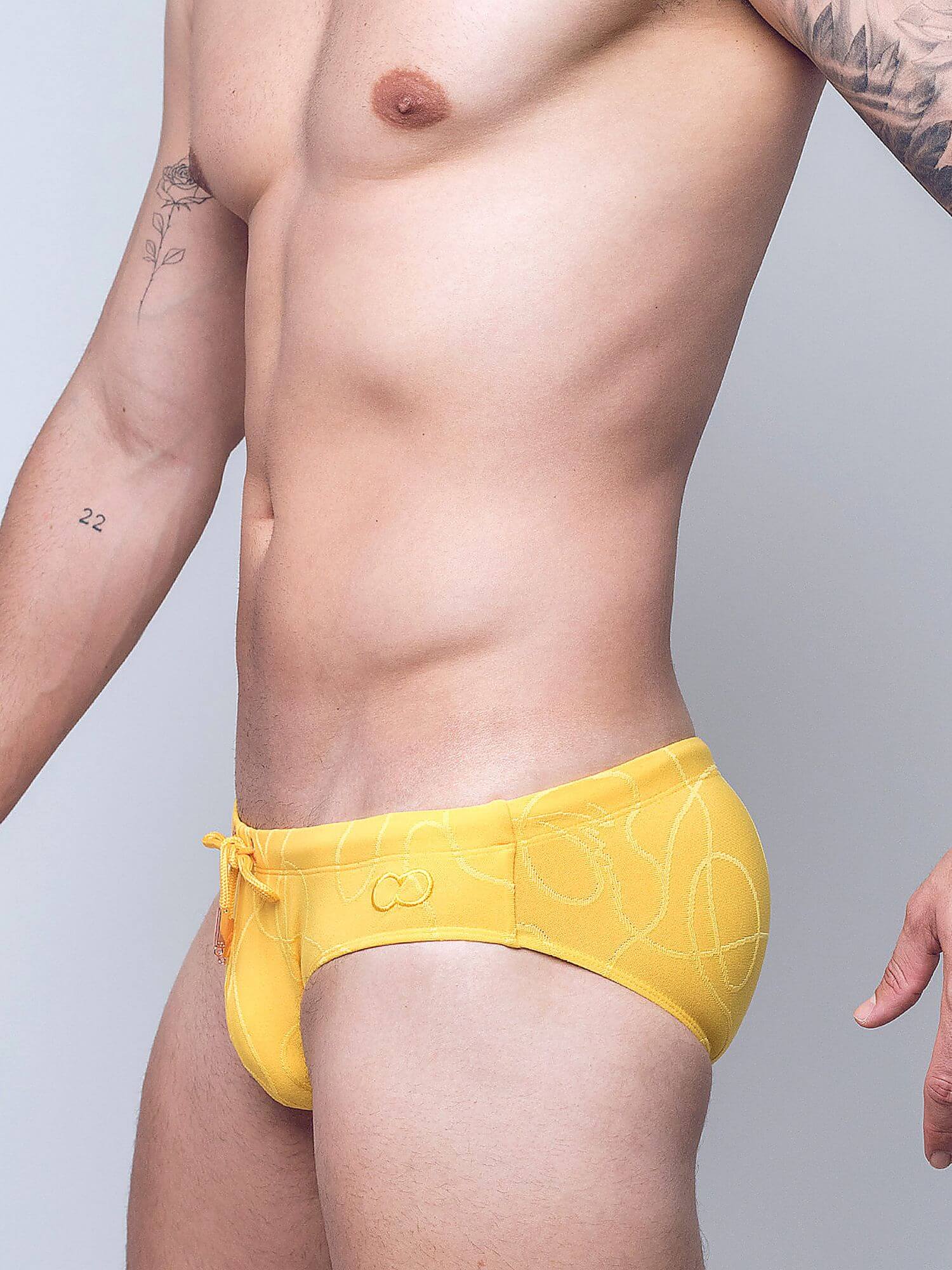A close-up of a man's lower torso wearing bright yellow swim briefs with a subtle white line pattern and a small embroidered logo on the front, against a light gray studio background.