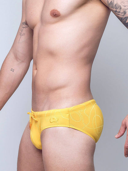 A close-up of a man's lower torso wearing bright yellow swim briefs with a subtle white line pattern and a small embroidered logo on the front, against a light gray studio background.
