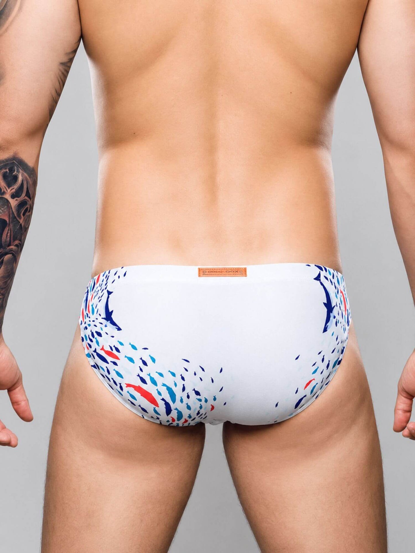 Rear view of a man wearing white swim briefs with a blue and red abstract pattern, set against a solid gray studio background.