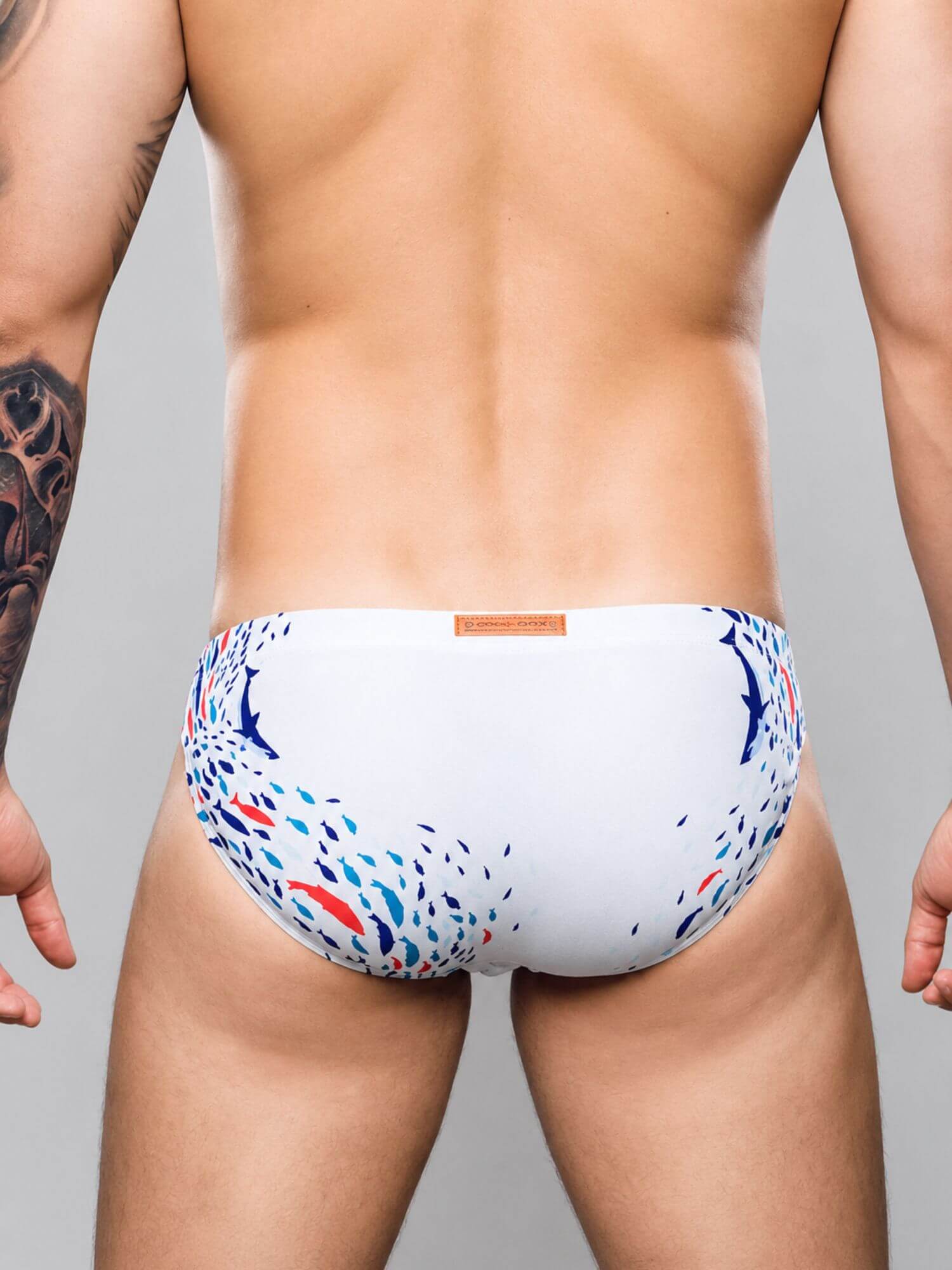 Rear view of a man wearing white swim briefs with a blue and red abstract pattern, set against a solid gray studio background.