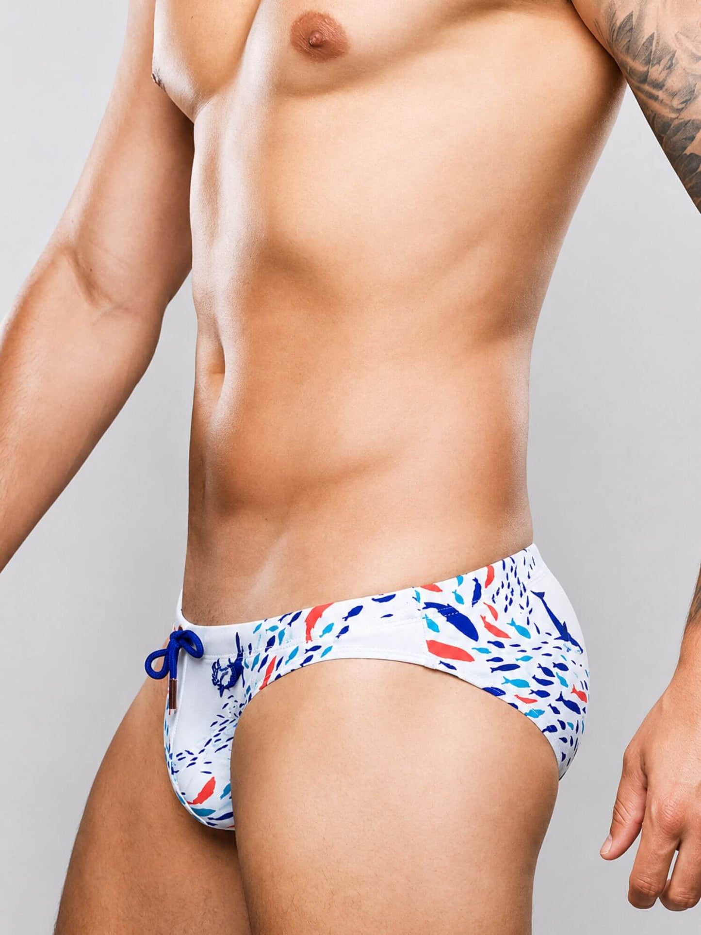 A close-up of a male model wearing low-rise swim briefs with a white base and a colorful abstract pattern of blue, red, and orange shapes, set against a plain gray studio background.