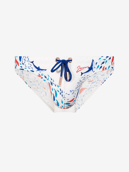 White bikini bottoms featuring a blue and red abstract print with shark motifs, a blue drawstring with gold-toned metal rings, and a red script logo on the right side.