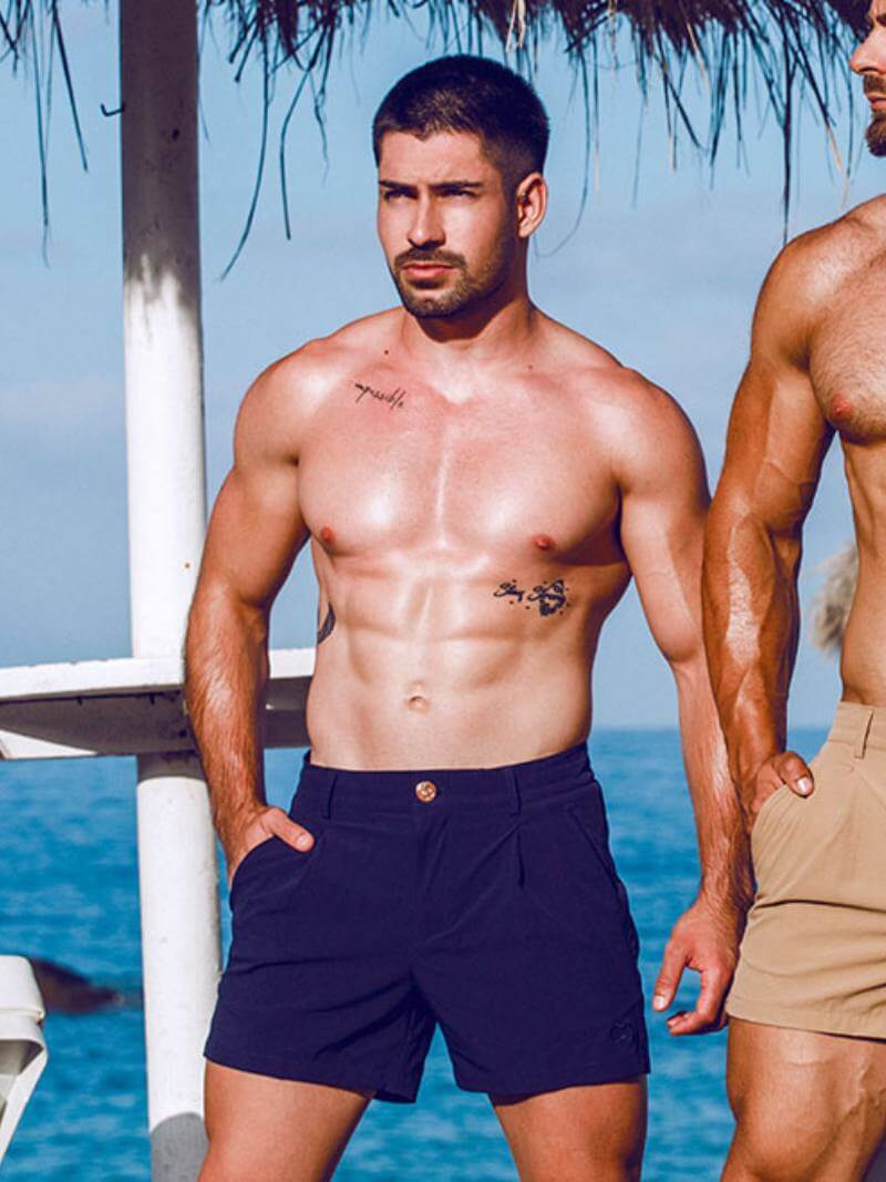 Two men standing by a body of water, one wearing navy shorts and the other in beige shorts.