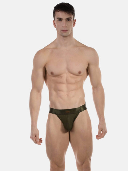 A muscular male model wearing a dark olive green jockstrap against a plain light gray studio background.