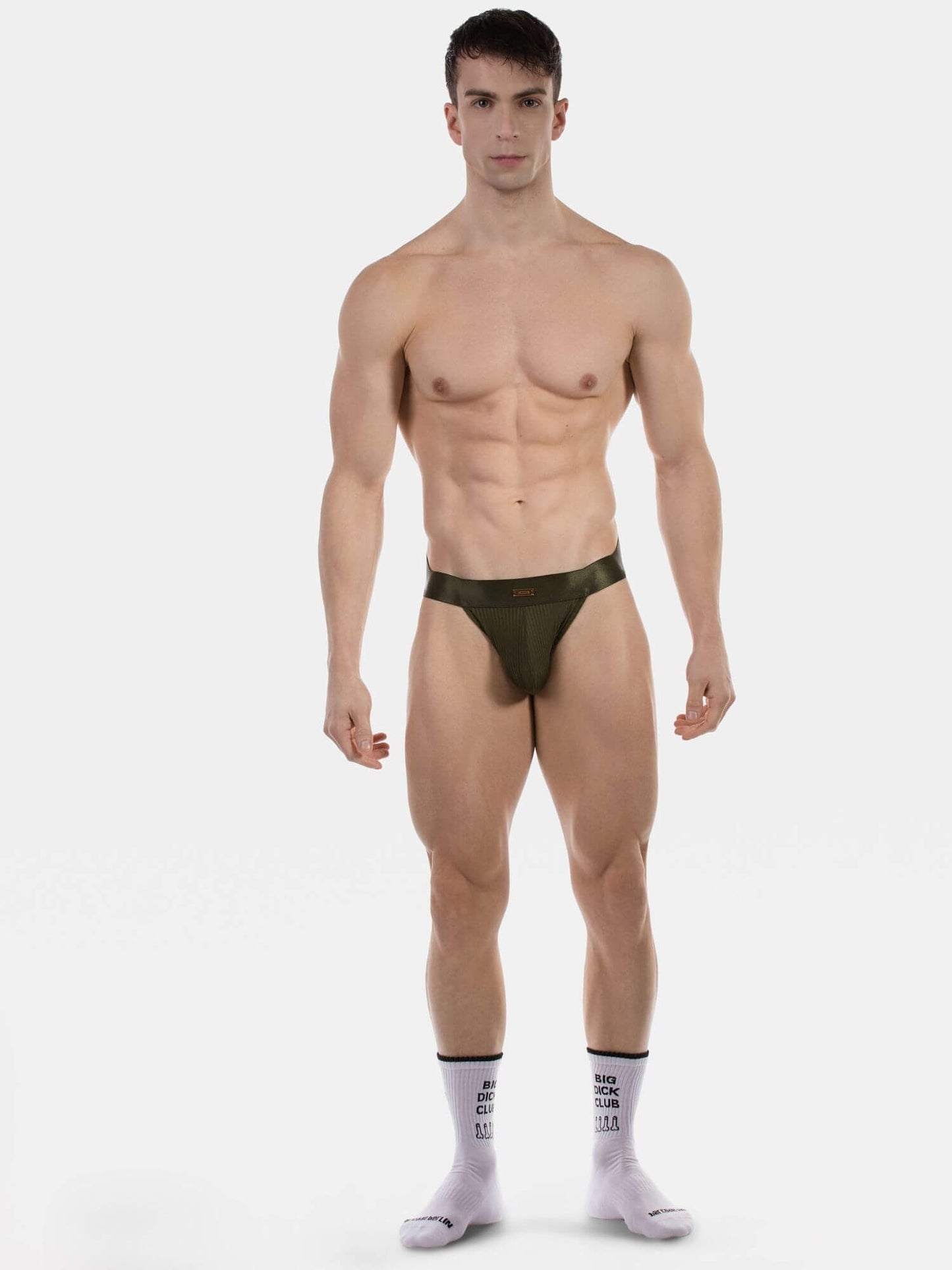 A muscular male model stands against a light gray studio background wearing a very minimal dark olive green jockstrap and light pink crew socks with black text.