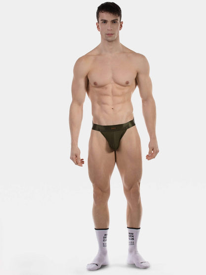 A muscular male model stands against a light gray studio background wearing a very minimal dark olive green jockstrap and light pink crew socks with black text.