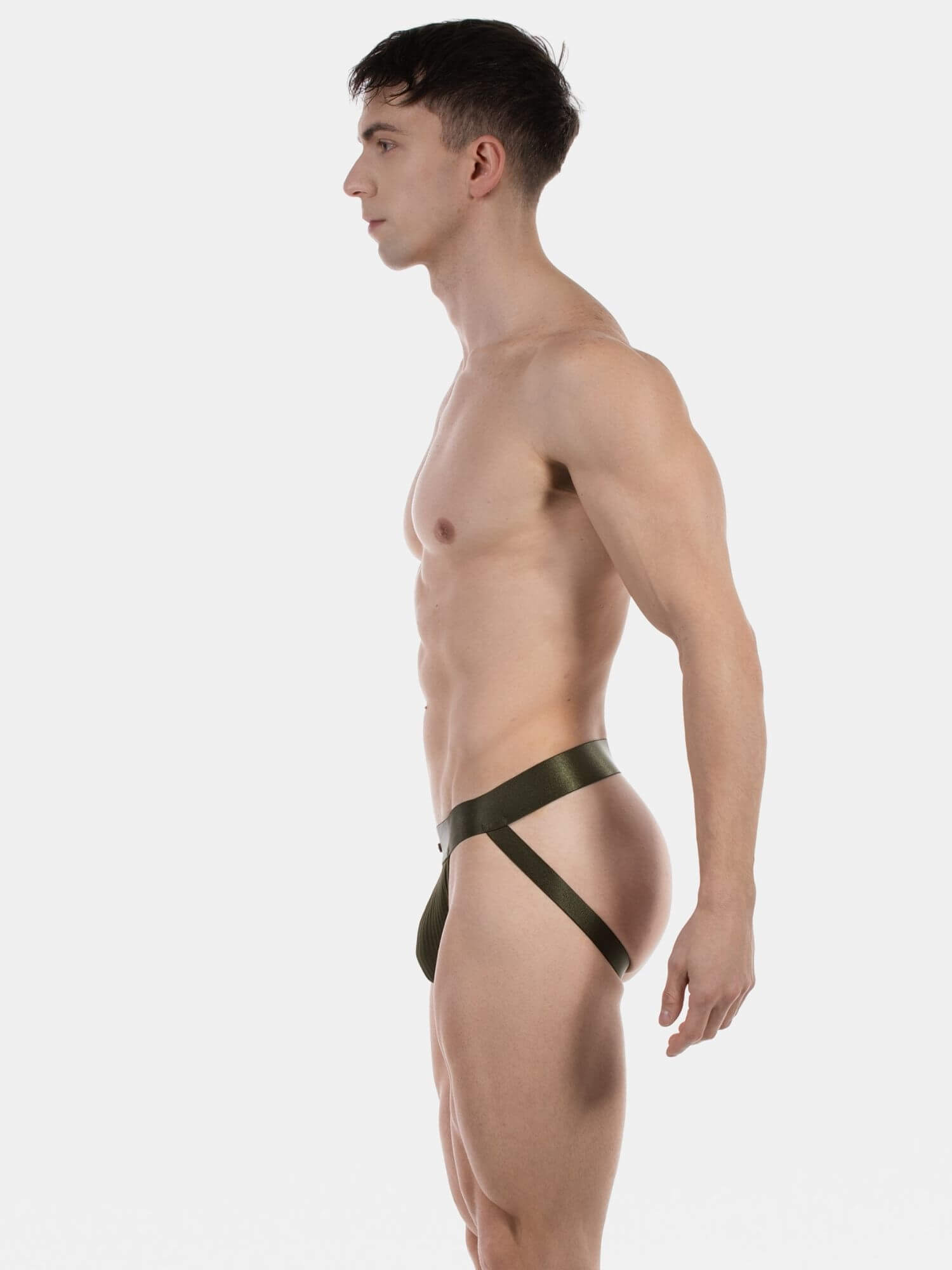 A muscular man in profile wearing a dark olive green minimal jockstrap underwear photographed against a seamless light gray studio background.