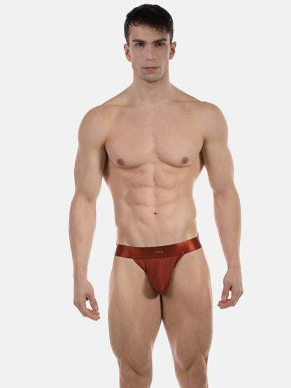 A muscular male model wearing a low-rise, satin-like reddish-brown jockstrap against a light gray studio background.