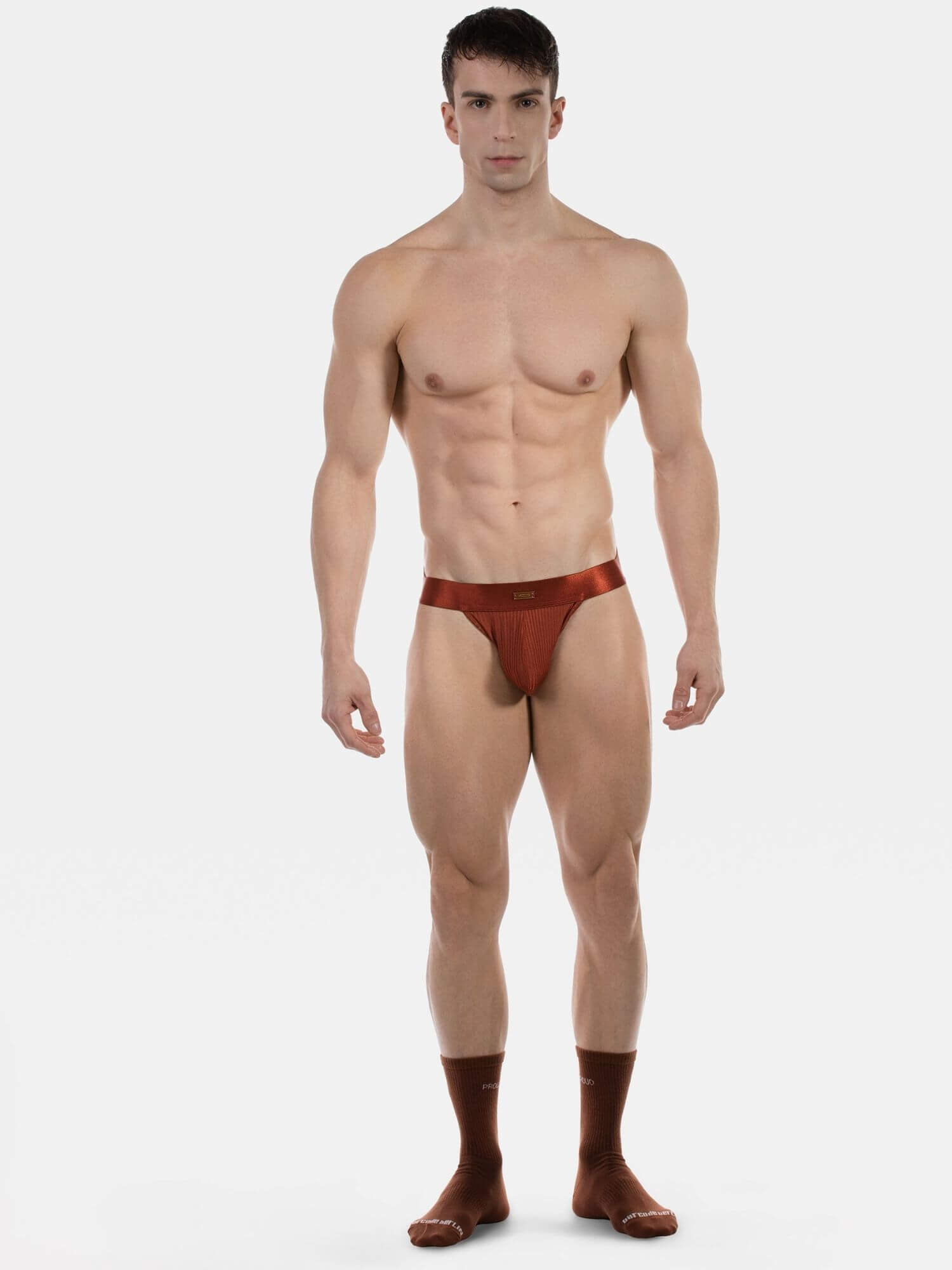 A muscular male model standing against a light gray studio background, wearing a very small brown jockstrap underwear bottom with matching brown socks.