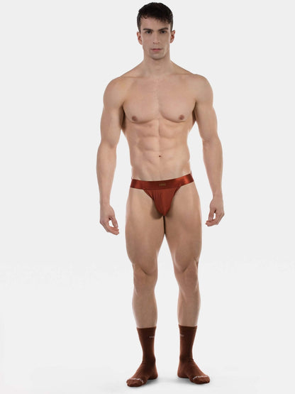 A muscular male model standing against a light gray studio background, wearing a very small brown jockstrap underwear bottom with matching brown socks.