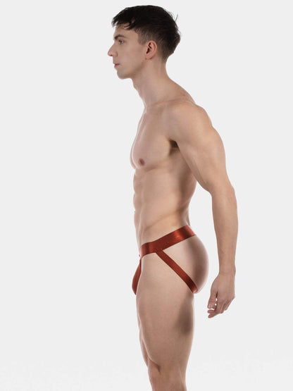 A muscular man in profile wearing a low-rise, satin-like red jockstrap with a wide waist strap, photographed against a seamless light gray studio background.
