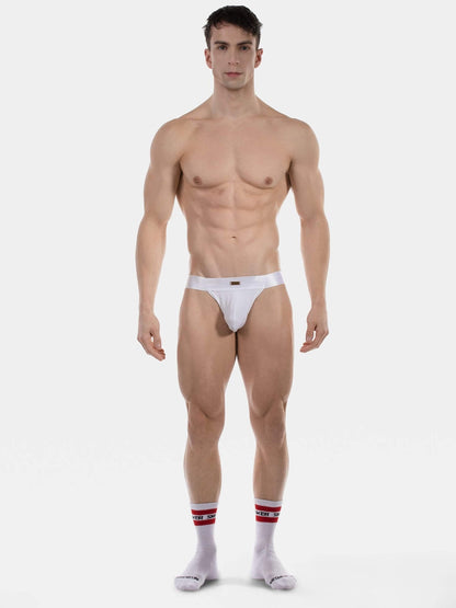 A muscular male model stands against a light gray studio background wearing minimal white briefs and white crew socks with red stripes.